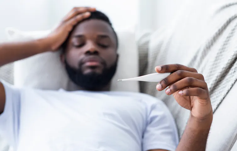man in bed takes own temperature and holds head