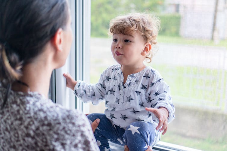 Toddler sat on window sill makes face at female carer