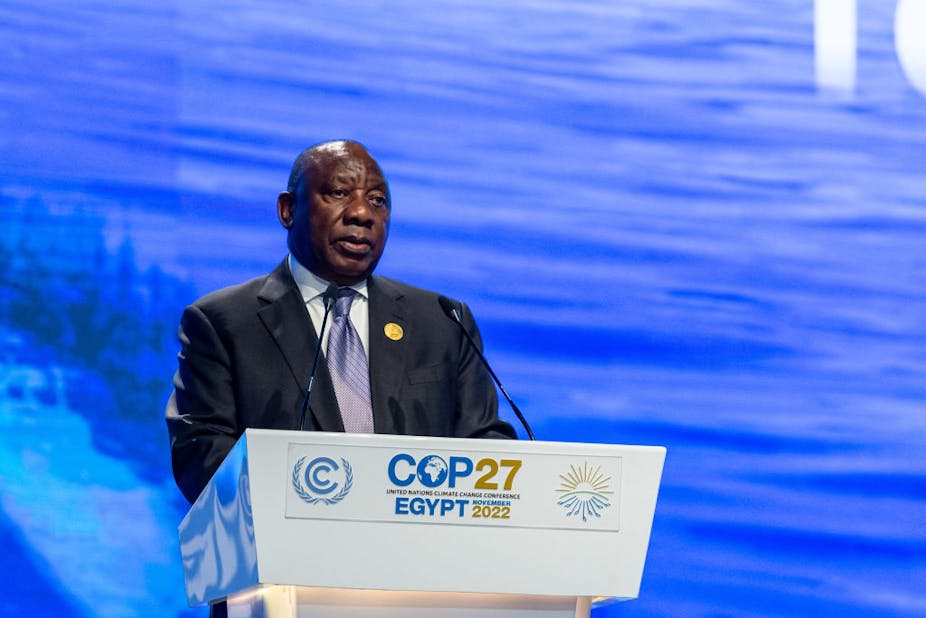 A man in a suit and tie stands behind a podium emblazoned with the words "COP27 Egypt 2022", against a blue backdrop