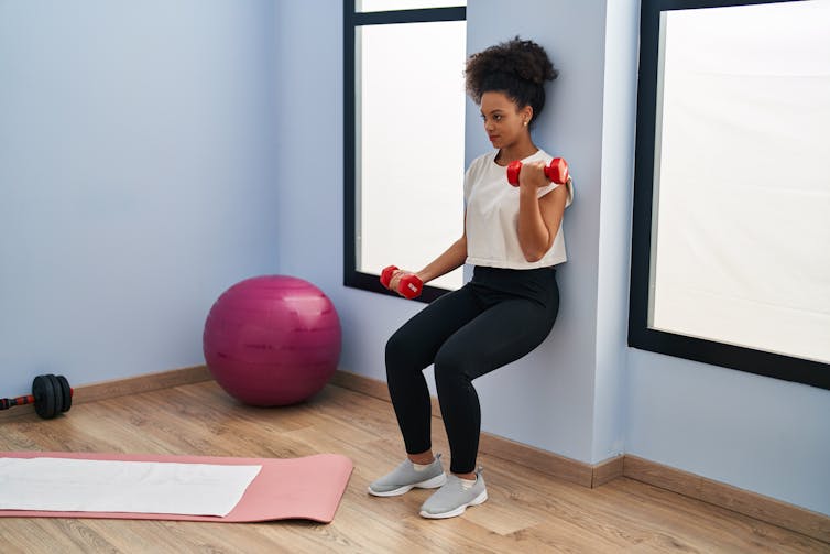 Young woman perform a wall sit while doing dumbbell bicep curls.