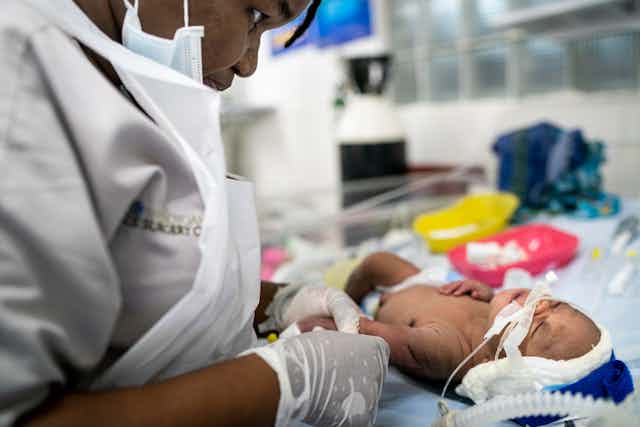 A woman dressed in white with a white apron and gloves leans over a baby and holds their arm. The baby has a diaper on and a ventilator tube coming out of their nose