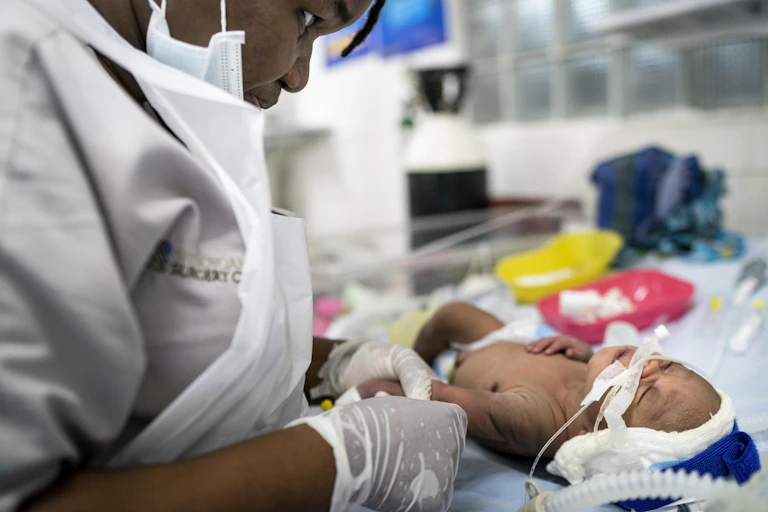 A woman dressed in white with a white apron and gloves leans over a baby and holds their arm. The baby has a diaper on and a ventilator tube coming out of their nose