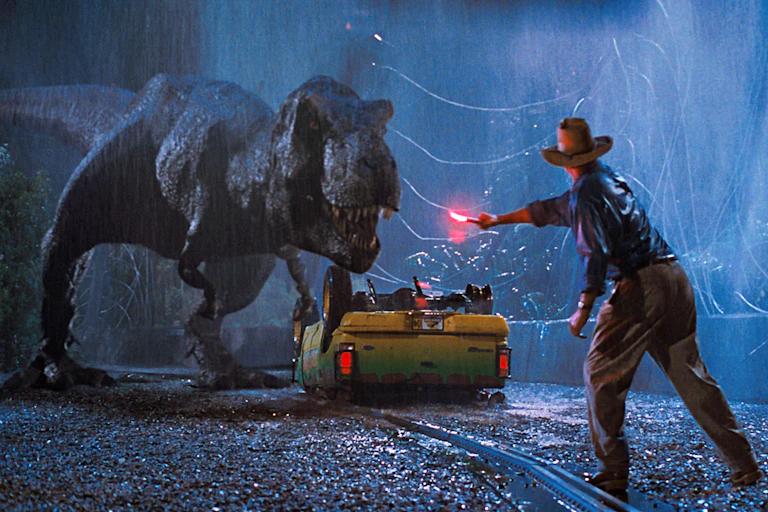 Dyn yn chwifio ffagl tra bod T. rex yn sefyll dros gar sydd wedi troi drosodd.