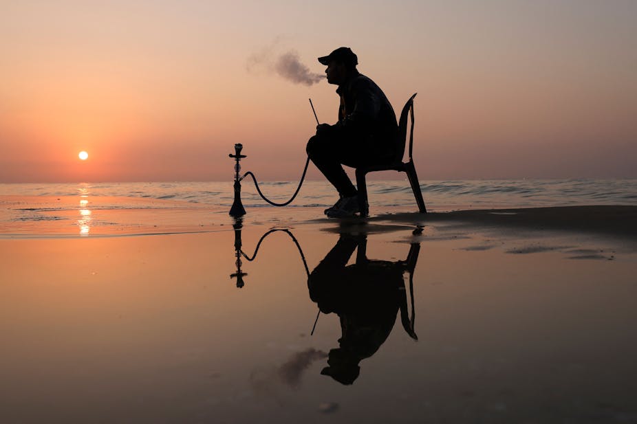 Un homme s'assoit sur une chaise au bord 'une plage en train de fumer de la chicha.