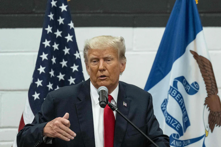 A man in a suit stands in front of several flags.
