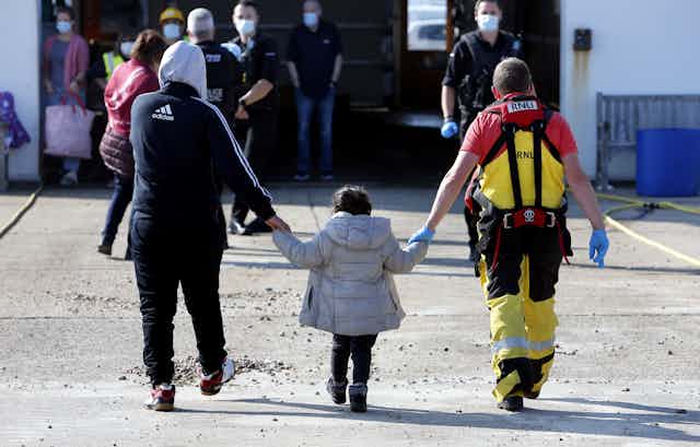 Photo from behind of an RNLI volunteer and an adult migrant each holding one hand of a child, who has arrived in the UK after being rescued from a small boat