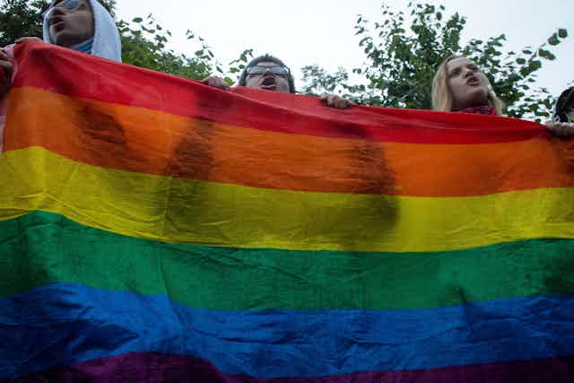 Three people stand behind a LGBT rainbow banner.