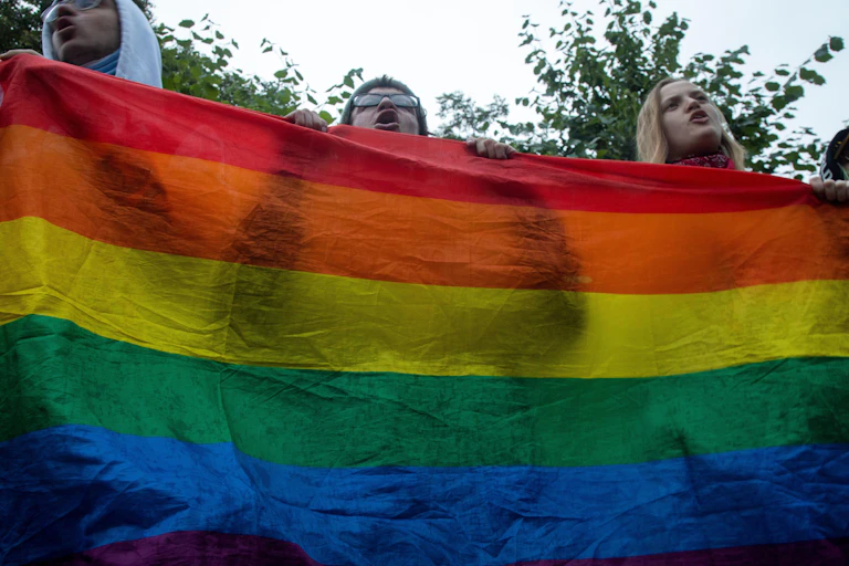 Three people stand behind a LGBT rainbow banner.