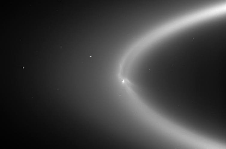 Against black space, a diffuse arc which is invisibly small icy particles scattering sunlight. A bright dot within the arc in Enceladus.