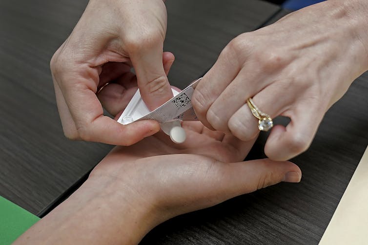A pair of hands pushes a white, round pill out of a blister packet into another person's waiting hand.