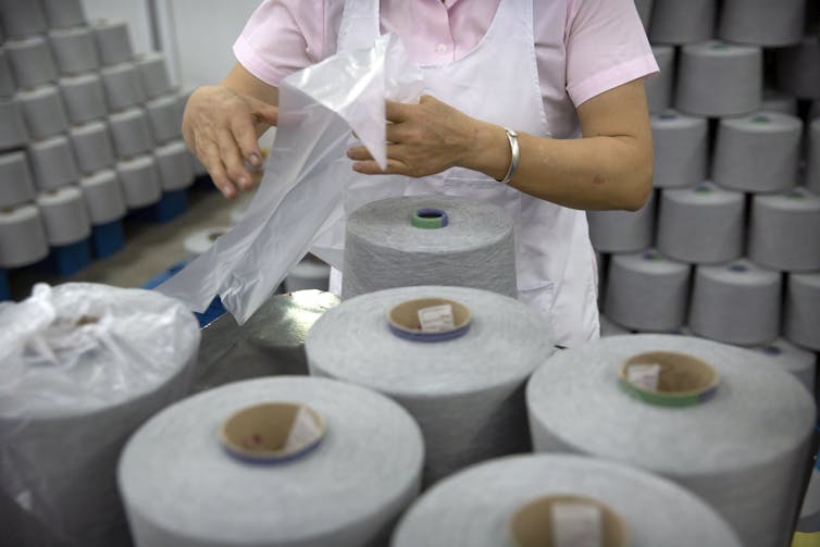 A person, seen from the shoulders down, opens a plastic bag over a massive spool of yarn sitting on a table. Yarn stacked in pyramids is seen in the background.