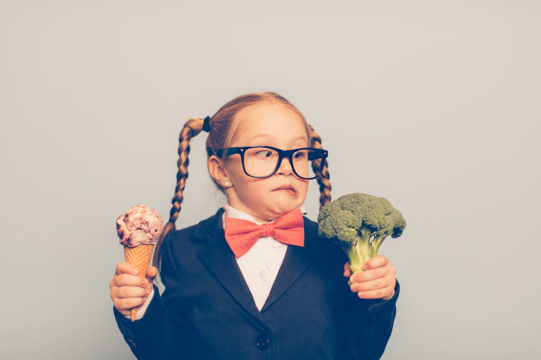 A young girl holds an ice cream cone in one hand and a stalk of broccoli in the other and looks askew at the broccoli.