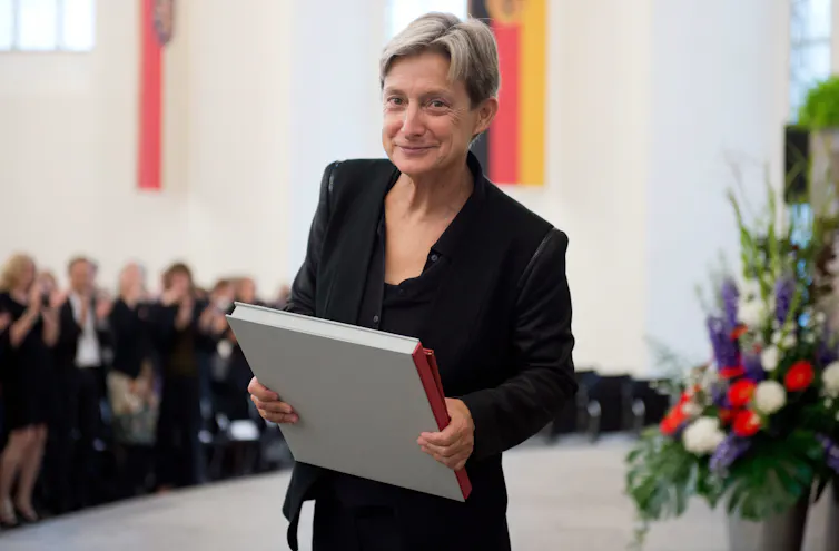A middle-aged individual in a black blazer and dress shirt smiles while holding a large hardcover book.