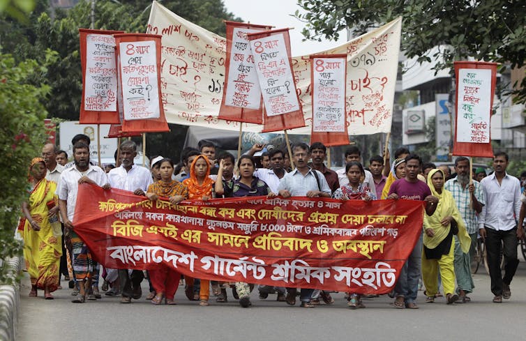 A group of people march down a street with protest signs and a large banner written in Bengali.