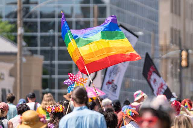 A rainbow flag waves above a crowd of people marching in a parade.