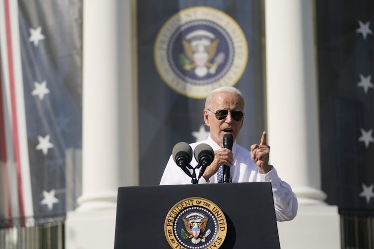 A man with white hair wearing aviator glasses speaks from behind a podium emblazoned with the crest of the U.S. president.