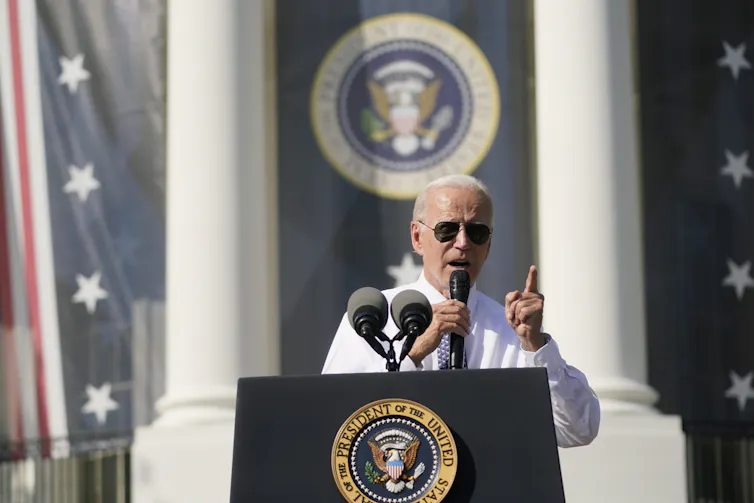 A man with white hair wearing aviator glasses speaks from behind a podium emblazoned with the crest of the U.S. president.
