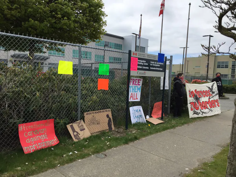 protest signs on a fence. People stand nearby holding a sign reading: no fences, no borders.