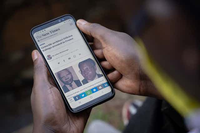 A pair of hands holding a mobile phone showing a news article by The New Times, with the headline 'Genocide architect Felcien Kabuga arrested in France'