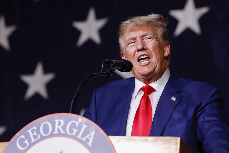 A middle-aged white man dressed in a business suit stands behind a podium and is speaking with his mouth wide-opened.