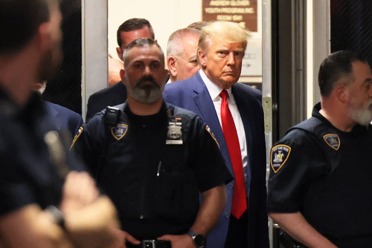 A middle aged white man dressed in a business suit is surrounded by court officers as he walks into a building.