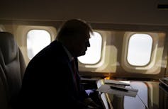 A man obscured in shadow, sitting in front of airplane windows.