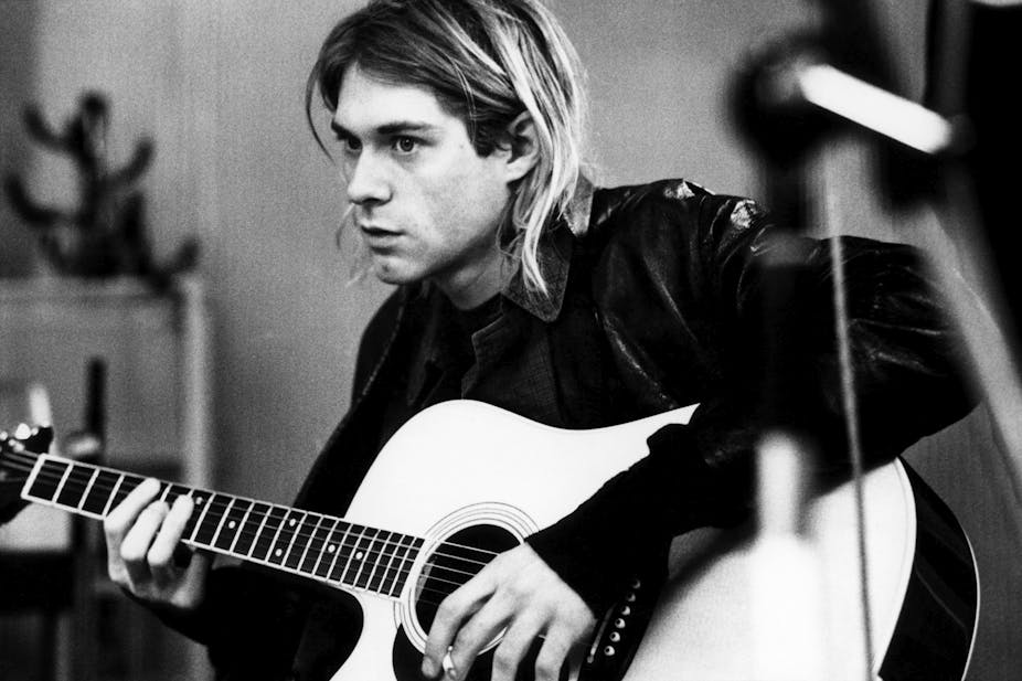 Kurt Cobain sat playing accoustic guitar in a leather jacket.