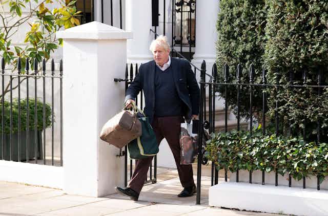 Boris Johnson carrying luggage out of a house through a gate.