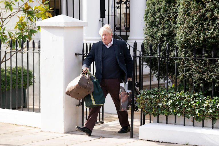 Boris Johnson carrying luggage out of a house through a gate.