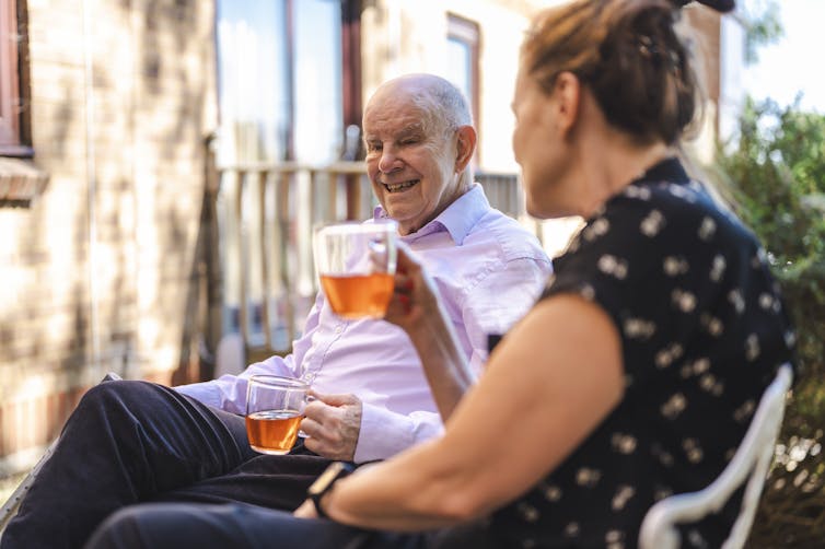 A middle aged woman and an older man sit side by side outdoors, smiling and drinking cups of tea together