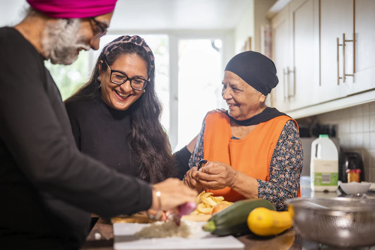 A young South Asian woman helps an older Sikh couple prepare food in a home kitchen