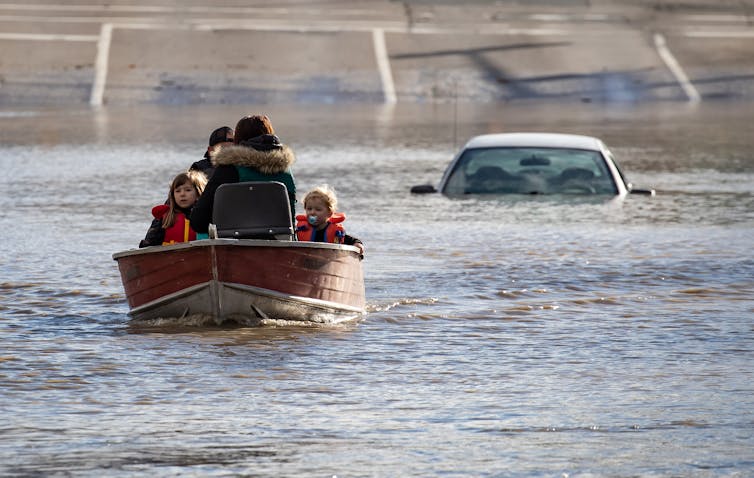 A woman and two children sit in a boat on the water. The roof of a car is seen beside them in the water.