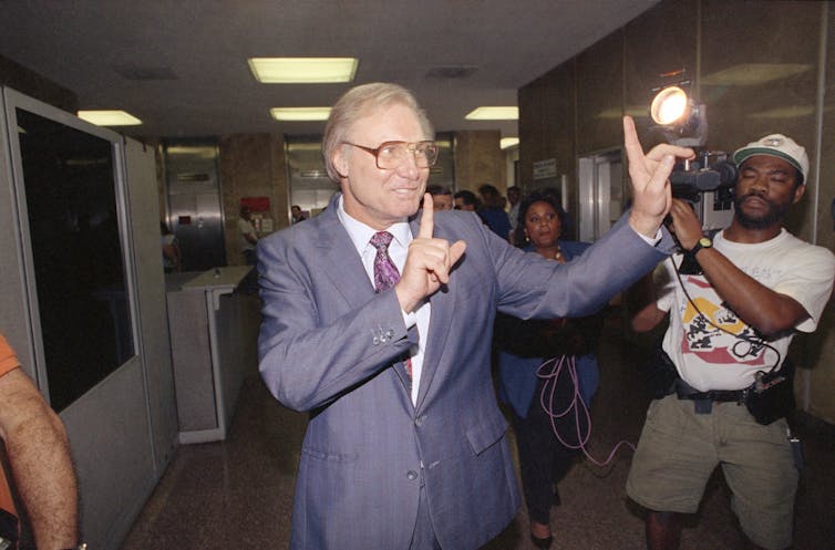 A man makes gestures while walking down a hallway. Behind him is a cameraman taking his pictures.