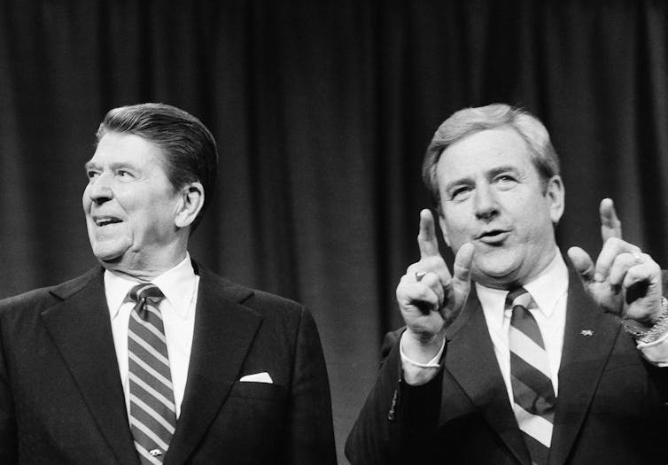 Two men in black suits sitting side by side and talking, and one of them on the right, making gestures.