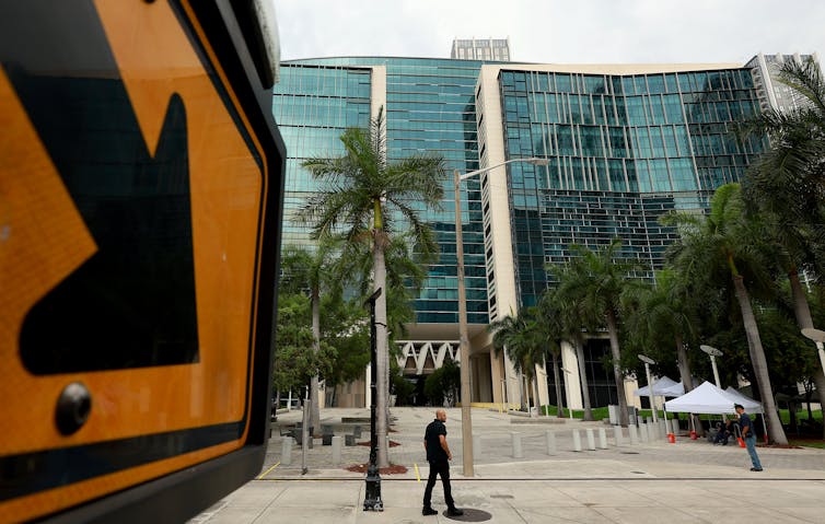 A yellow sign with a black arrow points toward a large glass building, with palm trees outside.