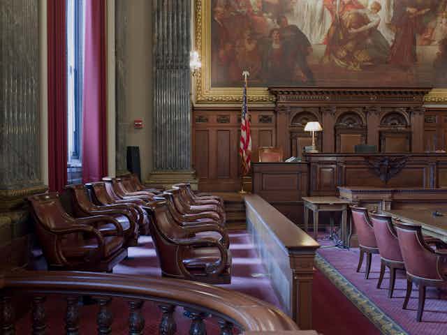 Three rows of seats with leather padding are seen in a formal looking room, with a U.S. flag behind and wooden panel.