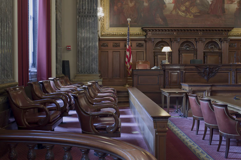 Three rows of seats with leather padding are seen in a formal looking room, with a U.S. flag behind and wooden panel.