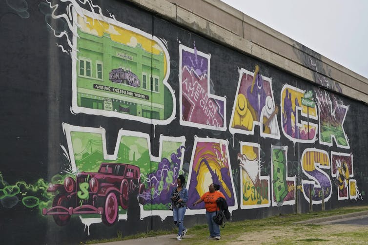 Two Black women walk past a brick wall with a mural painted on it that says 'Black Wall St.