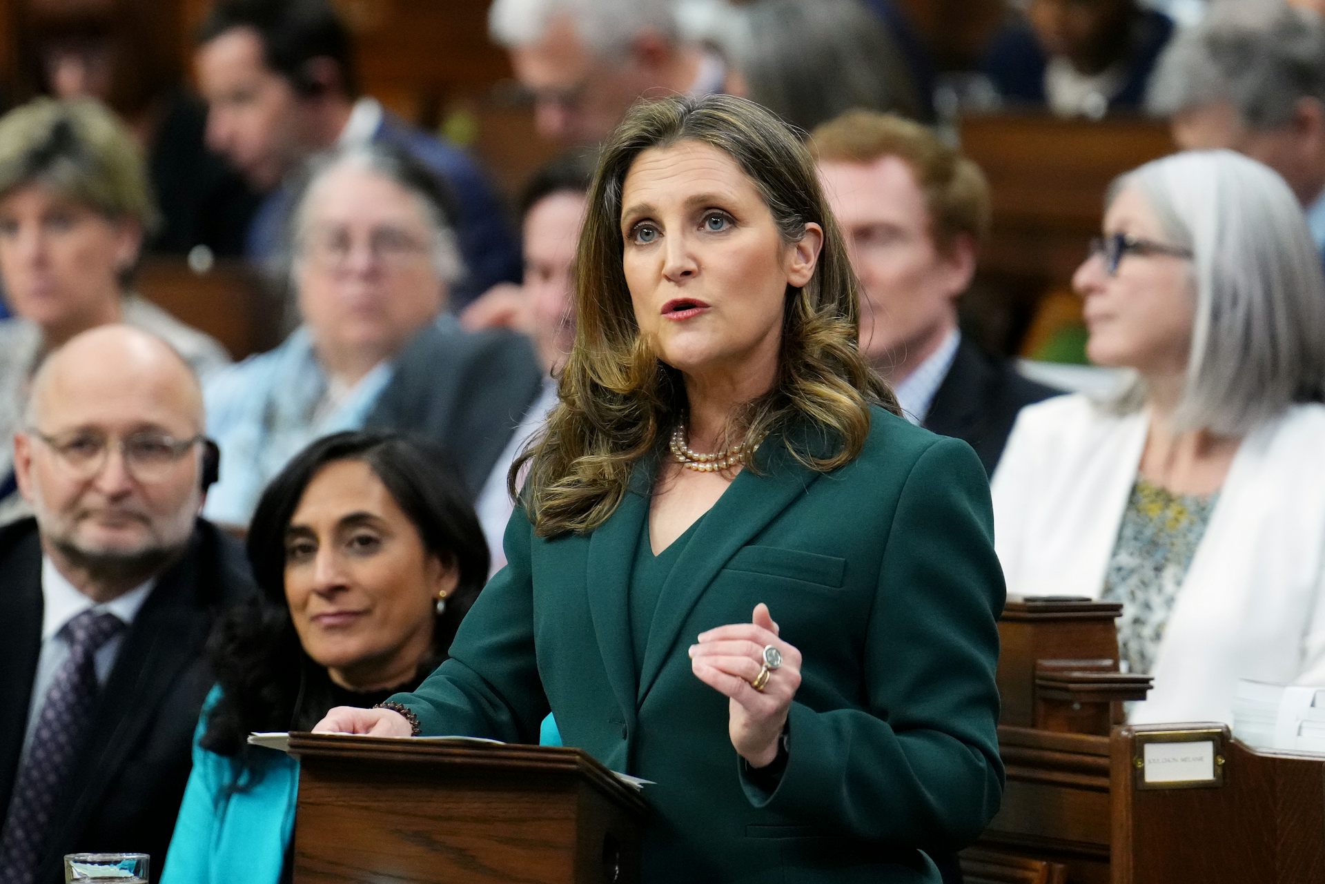A woman in a dark green suit gestures while speaking to a large crowd of people from behind a podium.