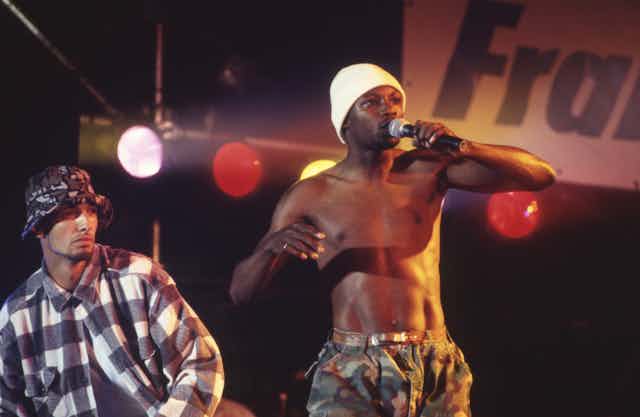 A shirtless man in a white hat sings into a microphone.