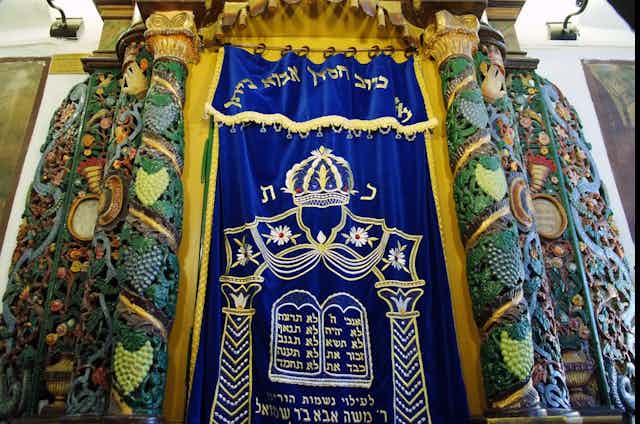 An intricately carved, colorful set of pillars on either side of an embroidered blue curtain with Hebrew phrases stitched into it.