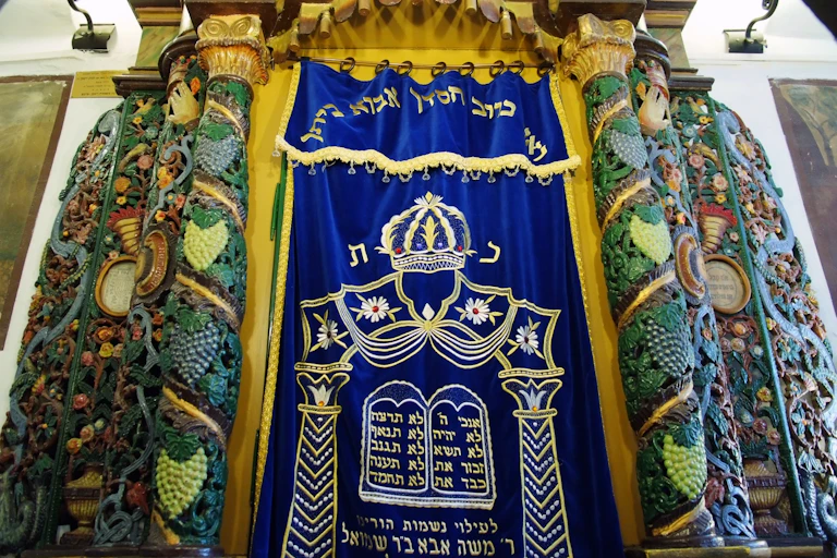 An intricately carved, colorful set of pillars on either side of an embroidered blue curtain with Hebrew phrases stitched into it.