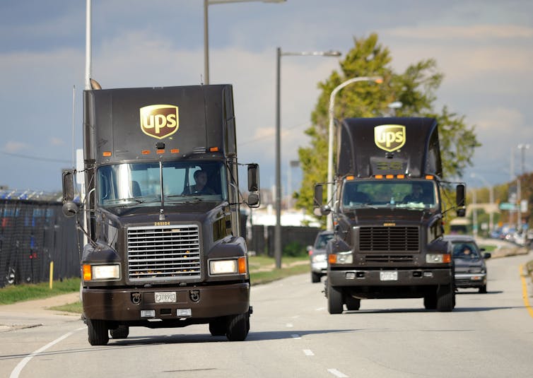 Two semi trucks with UPS logos drive down a highway toward the camera.