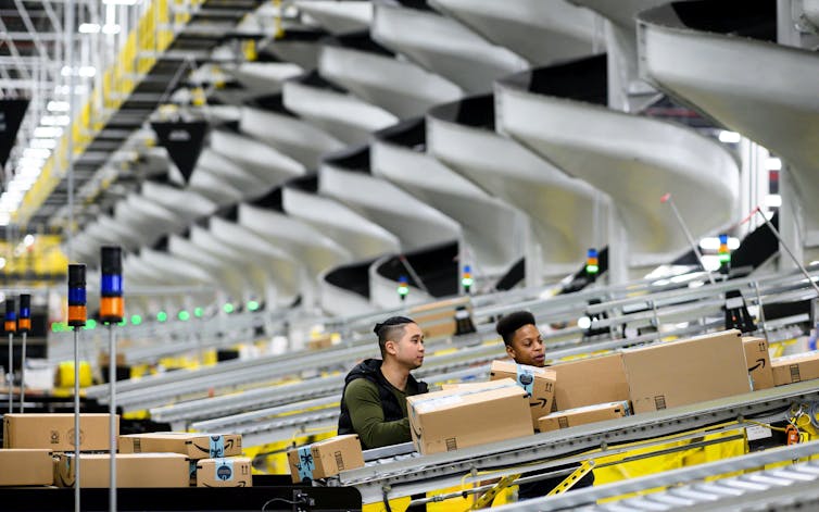 Two young men stand beside a conveyor belt with boxes on it. Several more belts just like it are behind them.