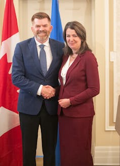 A woman in a burgundy suit shakes hands with a bearded man. Both smile at the camera.