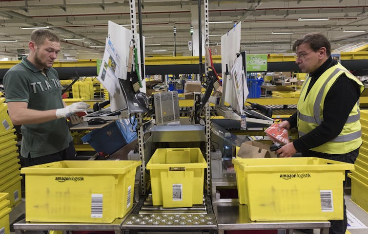 Two men holding products and scanners stand at computer stations with large bins beside them and stacks of bins in the background.