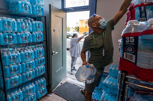A man in a t-shirt and shorts reaches for water bottles in a store room filled with cases of water.