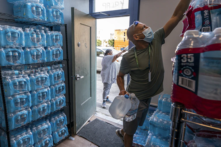 A man in a t-shirt and shorts reaches for water bottles in a store room filled with cases of water.