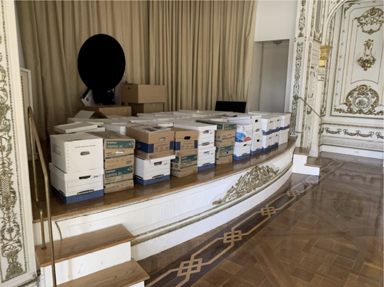 White and brown lidded boxes sit on top of white stage that has gold ornamentation. Gold curtains hang in the back of the stage.