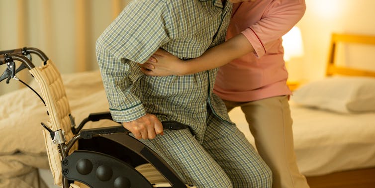 Care worker helping man from wheelchair to bed.